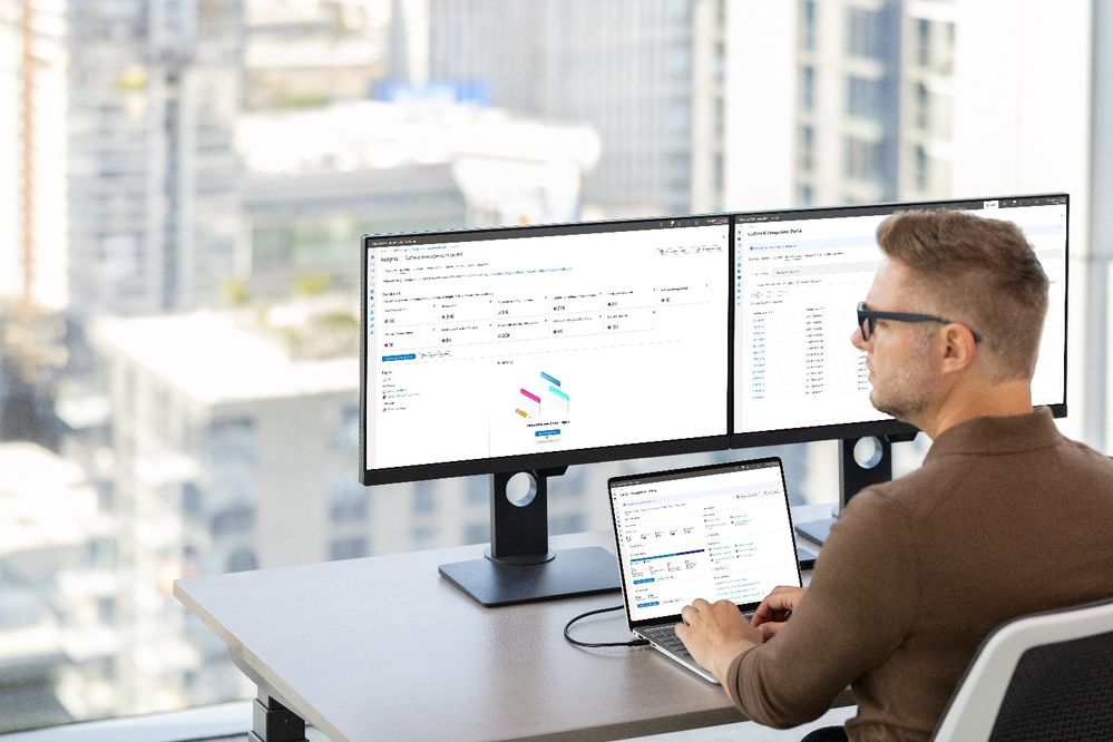 Person working at a desk with a Surface Laptop connected to dual external monitors, displaying productivity and data visualization tools in a modern office with a cityscape view.