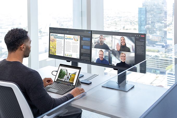 Person using a Surface Pro connected to dual external monitors, participating in a video conference on one screen while working on a document on the other, set in a modern office with a cityscape view